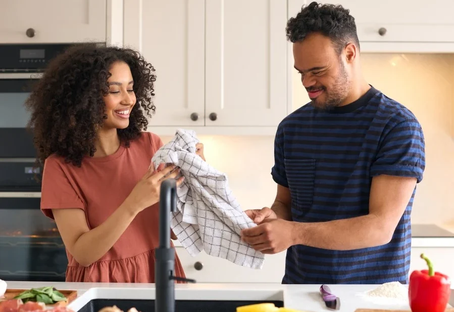 Disabled man with down syndrome in kitchen drying hands on tea towel with support worker